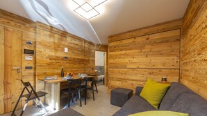A modern interior with wooden panel walls, a grey sofa with a green pillow, a dining area with a table and chairs, and a small kitchen area. - Marlies Ski in - Ski out 50m, Livigno, Italy (Livigno)