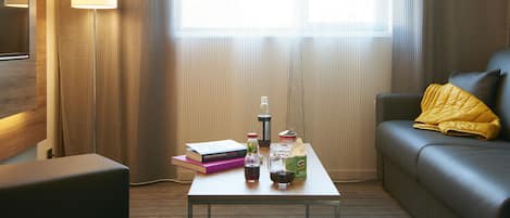 A hotel room with a sofa, a coffee table with books, a lamp, and a window with curtains.