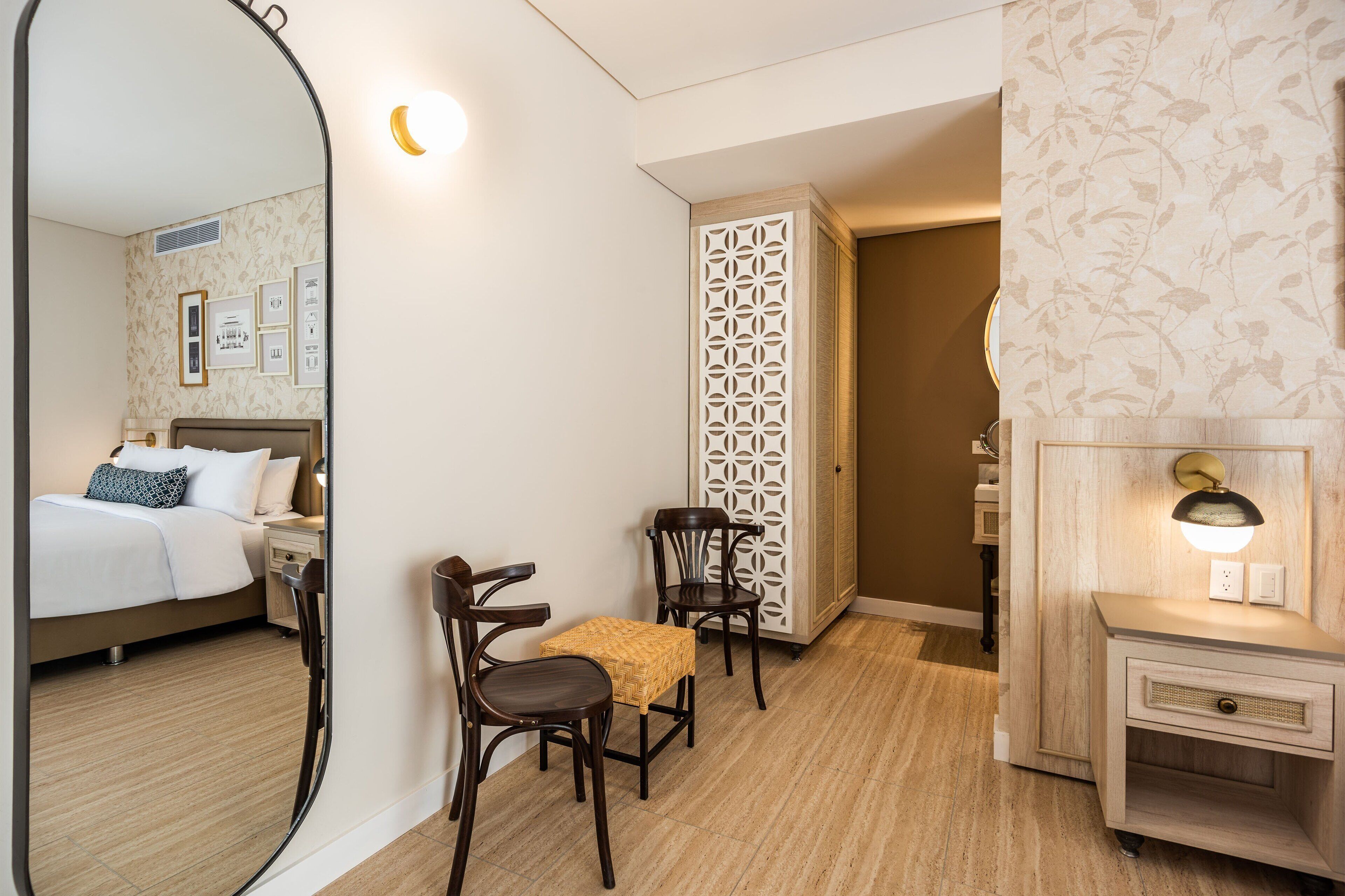 Quarto de hotel com cama, duas cadeiras, uma mesa pequena, um painel decorativo, uma mesinha de cabeceira e uma luminária fixada na parede.