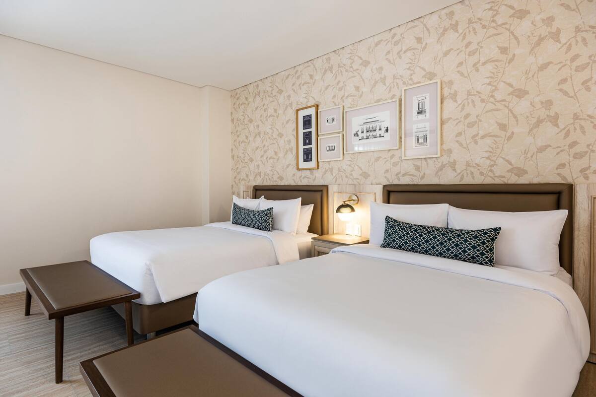 A hotel room with two beds, a nightstand, a small table, and framed pictures on the wall.