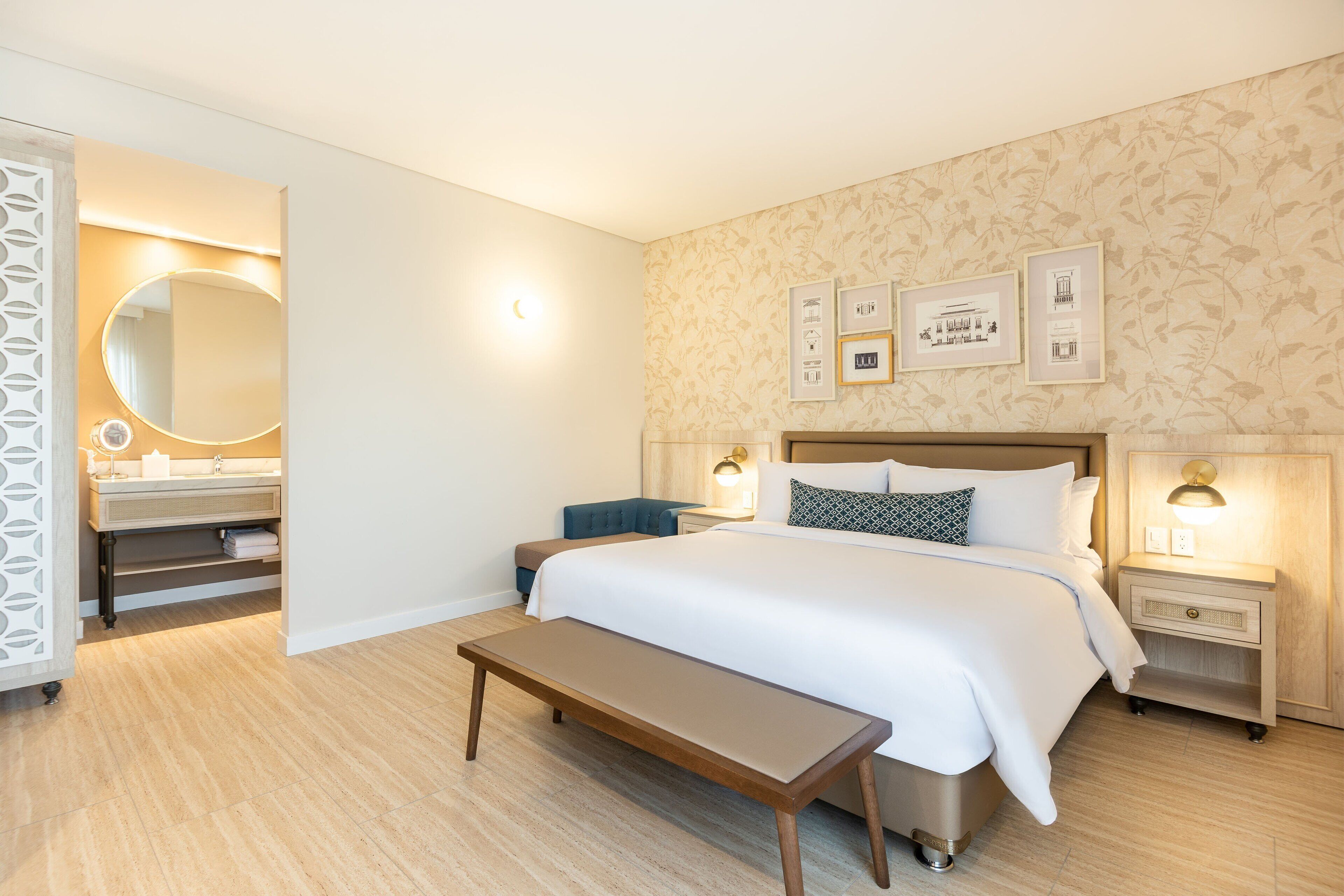Une chambre d’hôtel avec un grand lit, des tables de chevet, un banc, une salle de bain avec un miroir et des tableaux encadrés au mur.