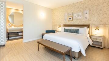 Habitación de hotel con una cama grande, mesitas de noche, un banco, un baño con espejo y cuadros en la pared.