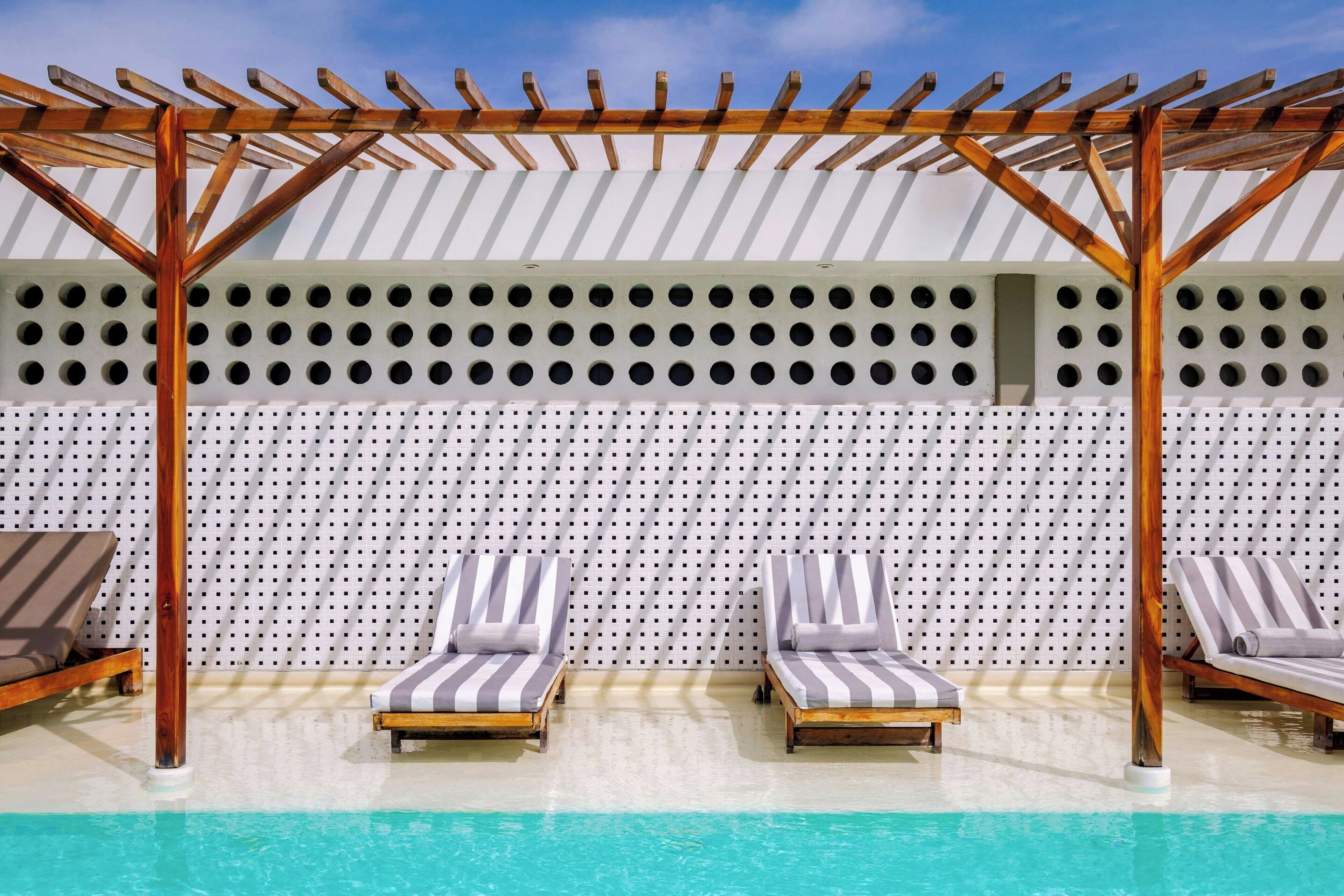 A poolside area with two sun loungers under a wooden pergola, a white perforated wall, and a clear blue pool.