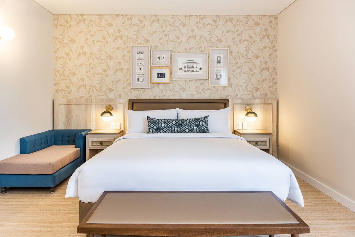 A hotel room with a large bed, bedside tables with lamps, a blue armchair, and a small bench.