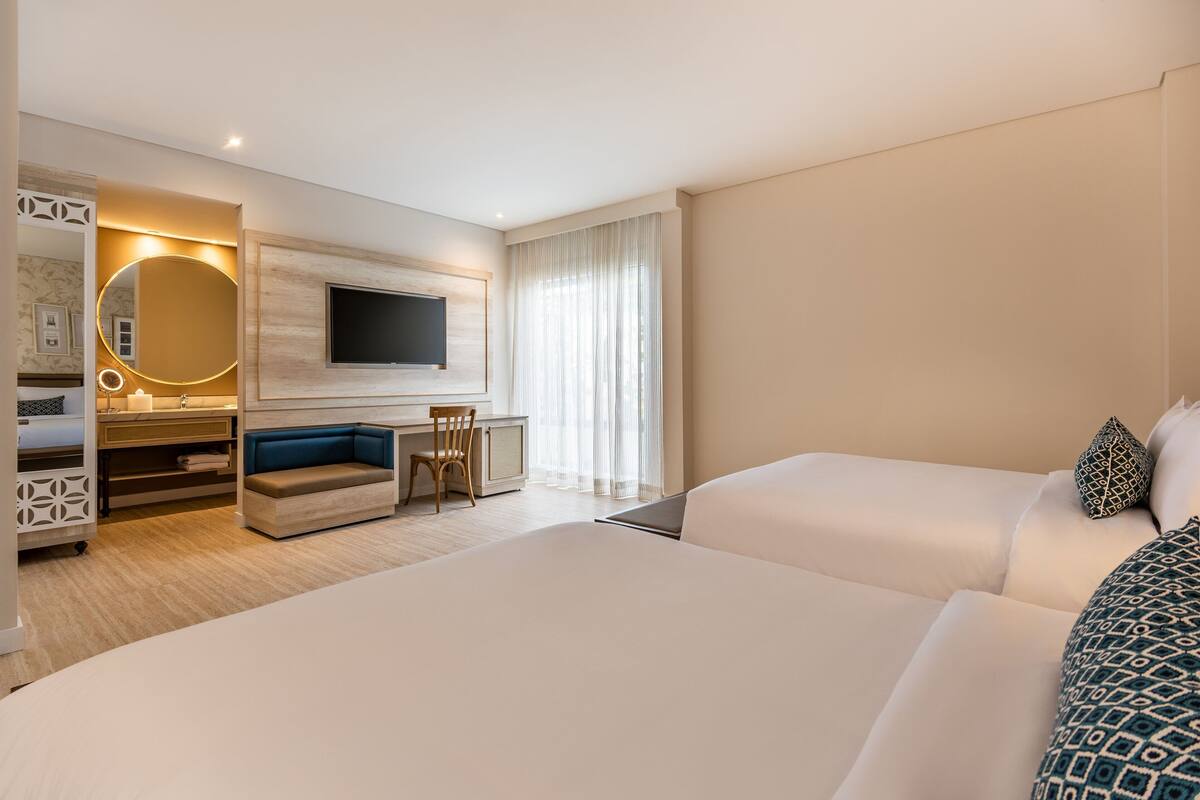 A hotel room with two beds, a desk with a chair, a TV, and a large window with curtains.