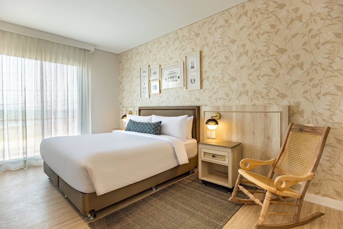 A neatly arranged bedroom with a large bed, a nightstand, a wooden rocking chair, and framed pictures on the wall.