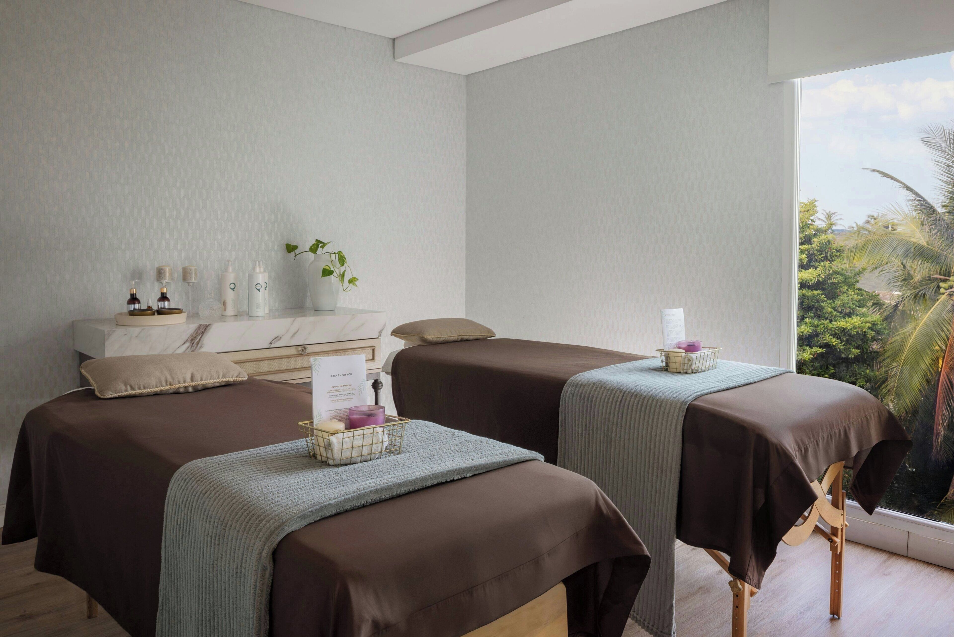 A spa room with two massage tables, a wooden bench, a small white cabinet, and a window with a view of greenery.