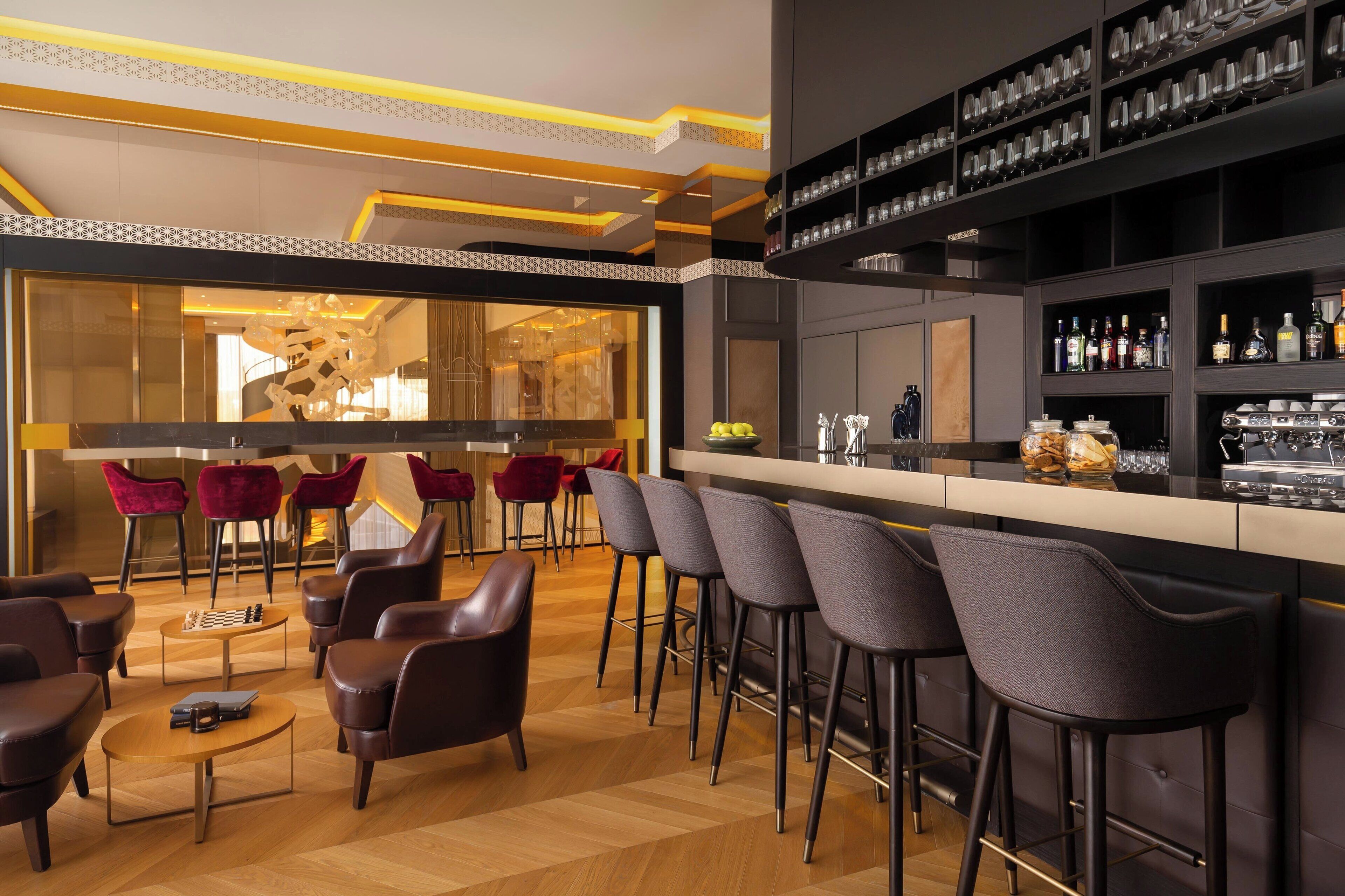 Uma área de bar moderna com banquetas altas, um tabuleiro de xadrez e um balcão repleto de garrafas e copos.