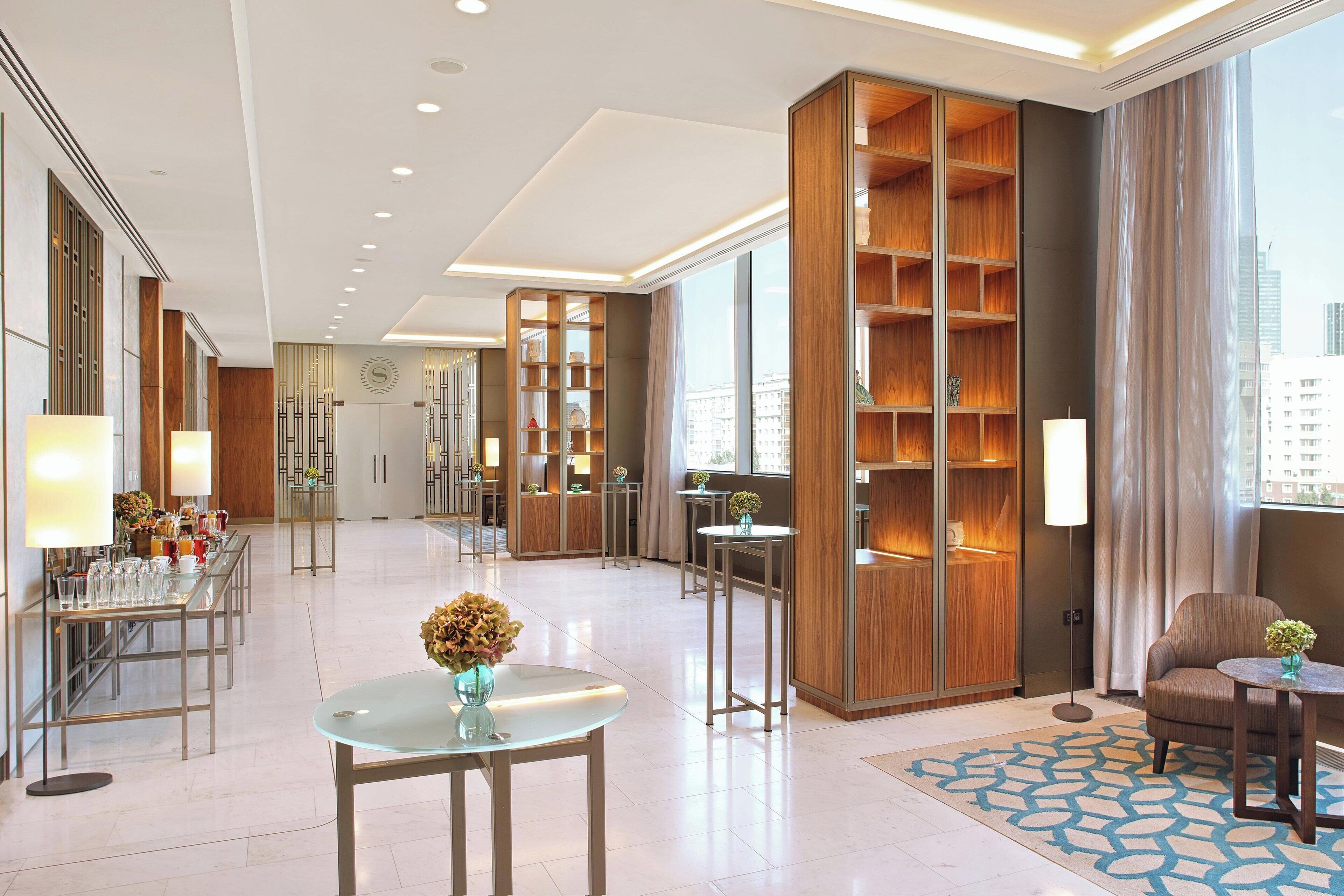 A spacious hotel lobby with a modern design, featuring a reception desk, seating areas, and a large bookshelf.