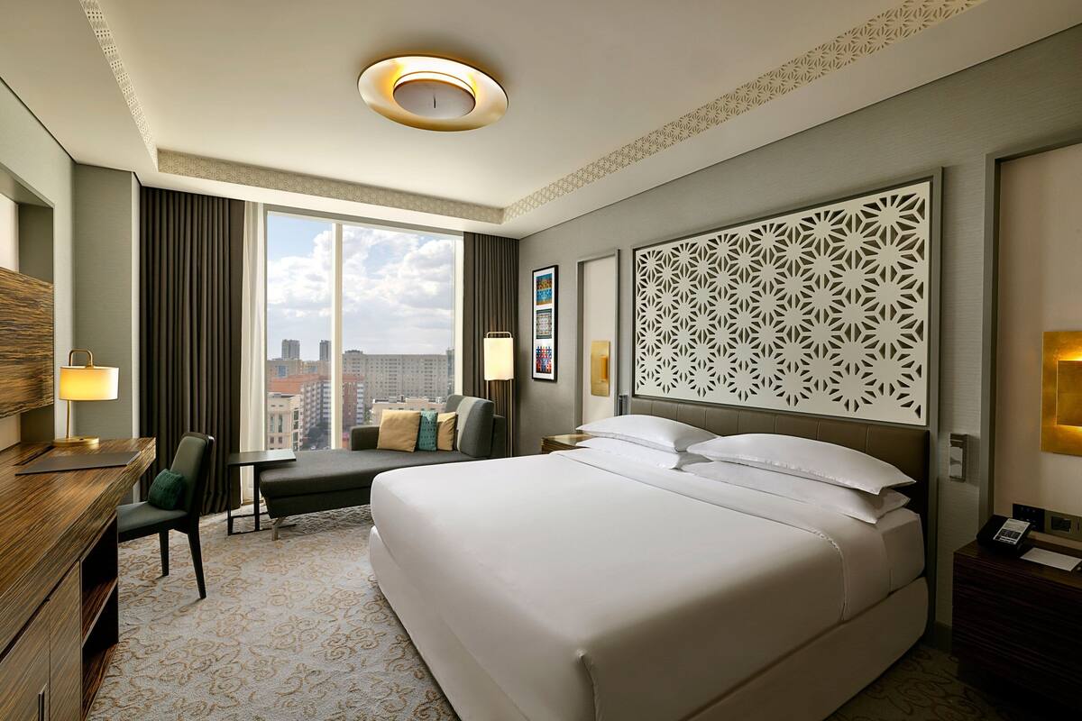 A hotel room with a large bed, a desk with a lamp, a chair, a sofa, a decorative wall panel, and a city view through the window.