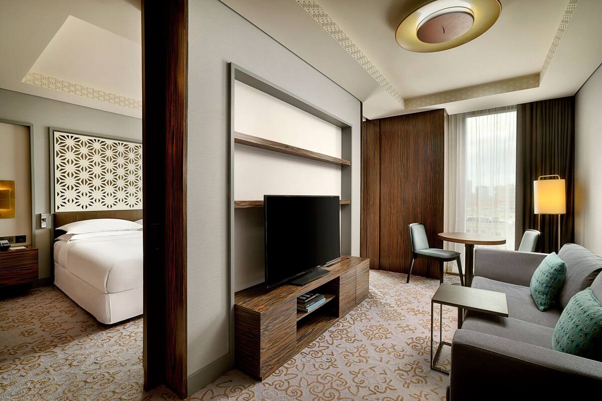 A hotel room with a bed, a TV on a wooden stand, a sofa, a small table, and a window with curtains.