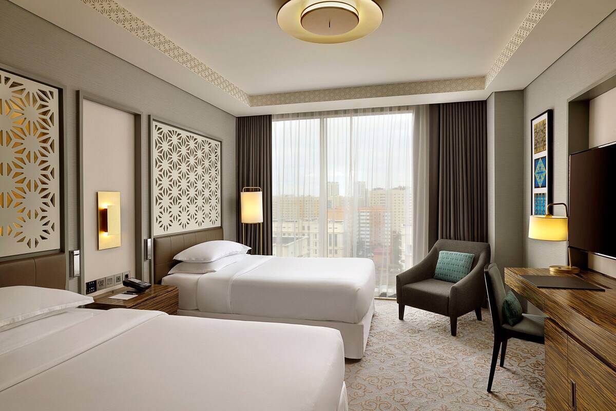 A hotel room with two beds, a desk, a chair, a TV, and a large window with curtains.