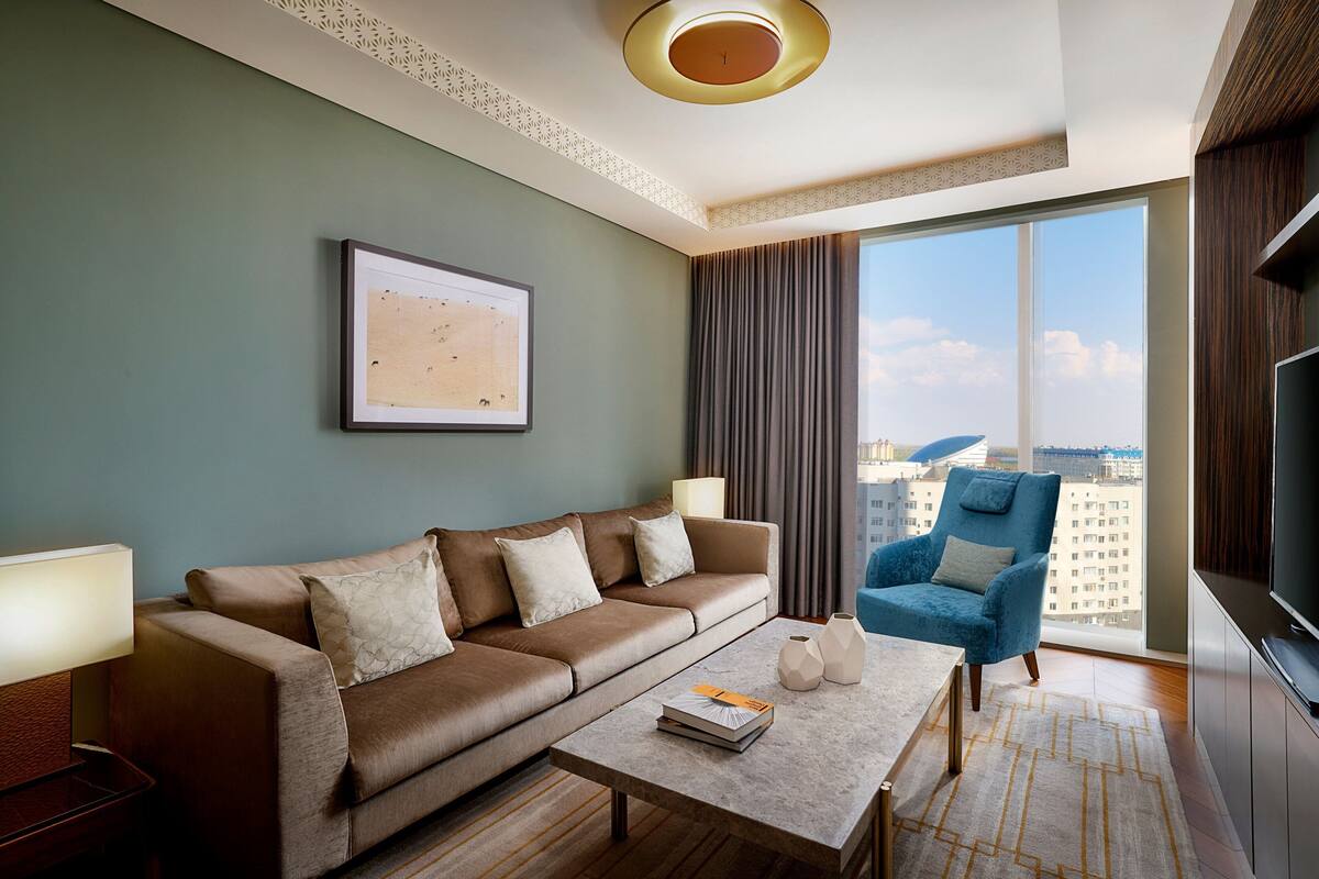 A modern hotel room with a leather sofa, a blue armchair, a flat-screen TV, and a large window offering a city view.