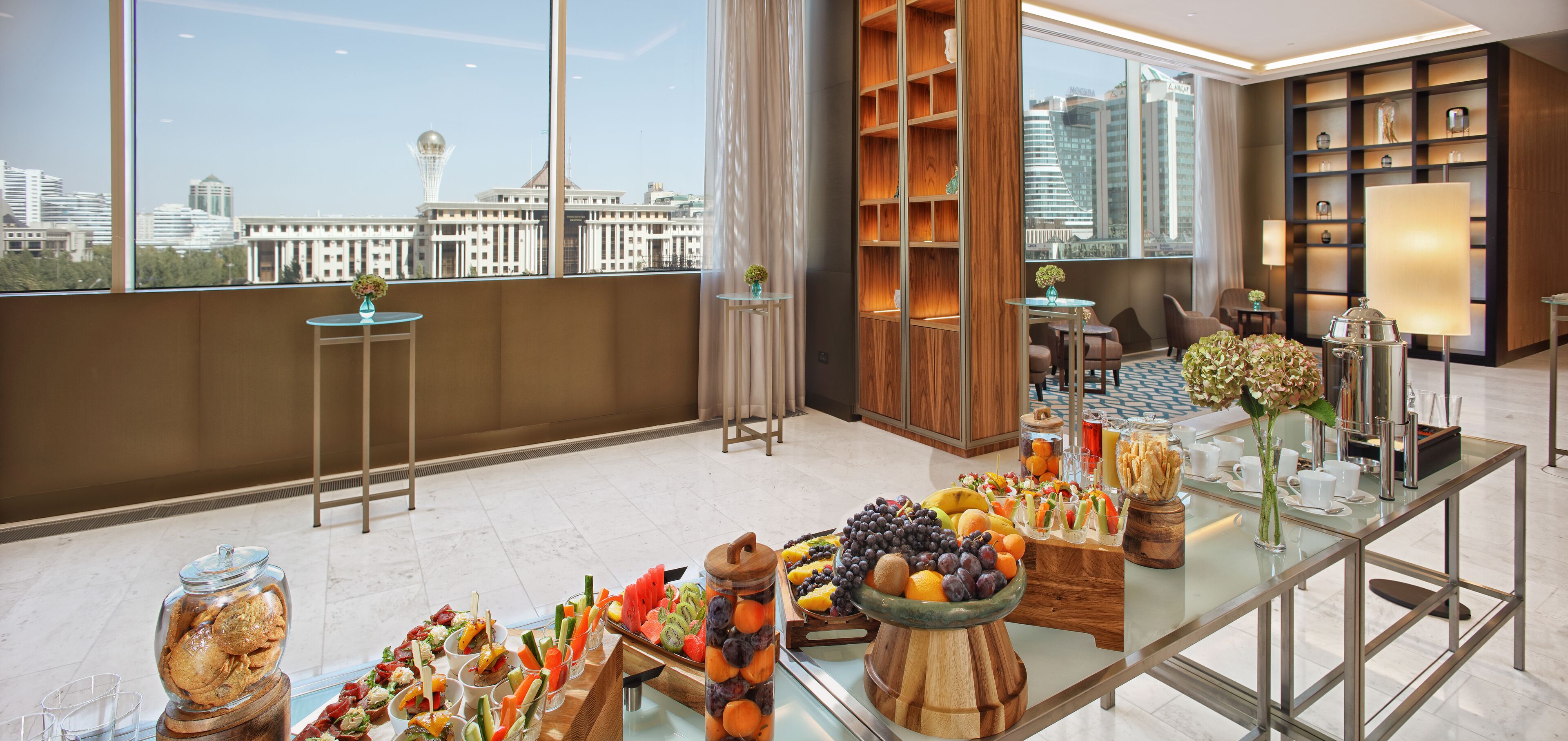 A modern hotel room with a city view, a buffet table with various fruits and snacks, a seating area with chairs, and a bookshelf with decorative items.