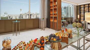 A modern hotel room with a city view, a buffet table with various fruits and snacks, a seating area with chairs, and a bookshelf with decorative items.