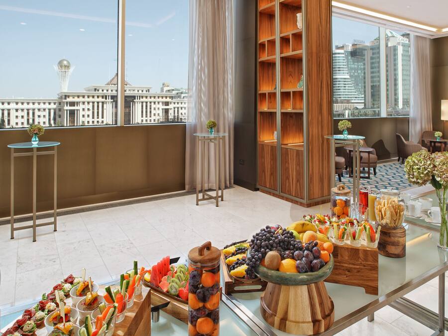 A modern hotel room with a city view, a buffet table with various fruits and snacks, a seating area with chairs, and a bookshelf with decorative items.