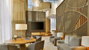 Un lobby moderno de hotel con una mesa de comedor redonda, sofás grises, un televisor de pantalla plana y una pared decorativa con líneas verticales.