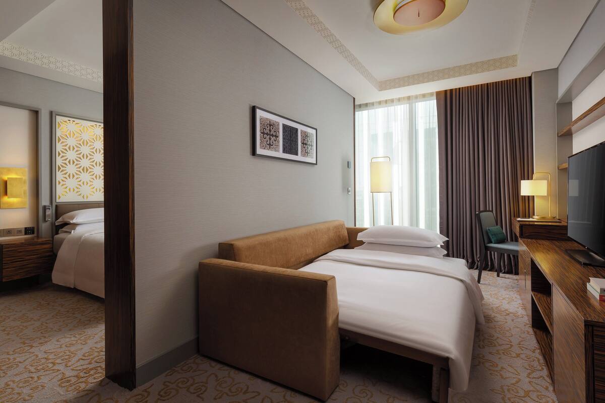 A hotel room with a large bed, a sofa, a desk with a TV, and a window with curtains.