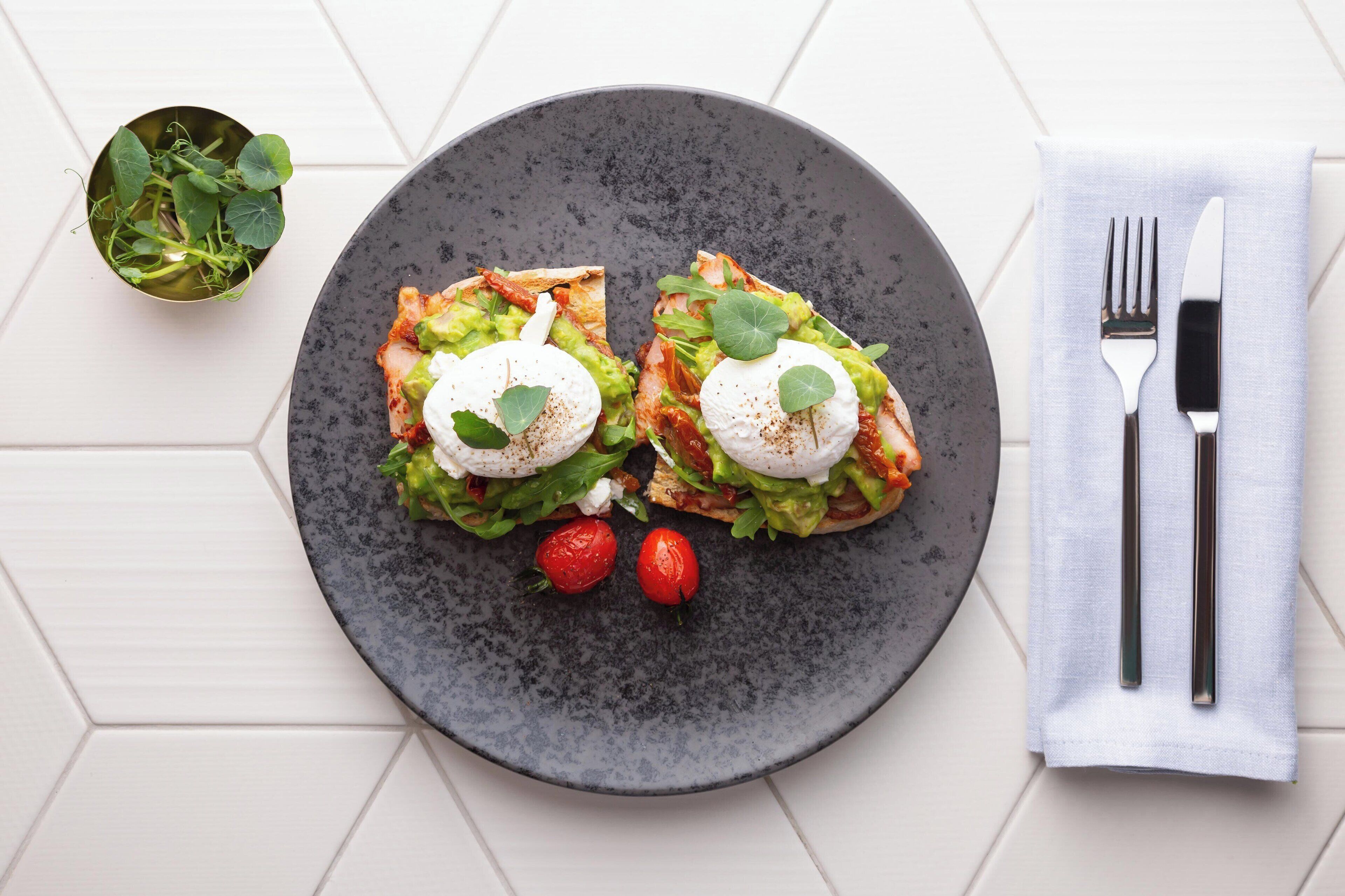 Un plato con dos sándwiches abiertos cubiertos de aguacate, hojas verdes y huevos pochados, acompañado de dos fresas y un pequeño bol de ensalada.
