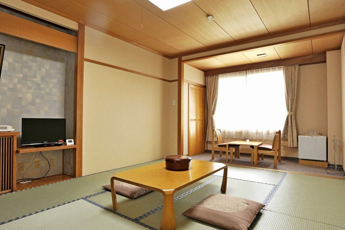 A traditional Japanese room with tatami flooring, a low table with cushions, a television, and a window with curtains.