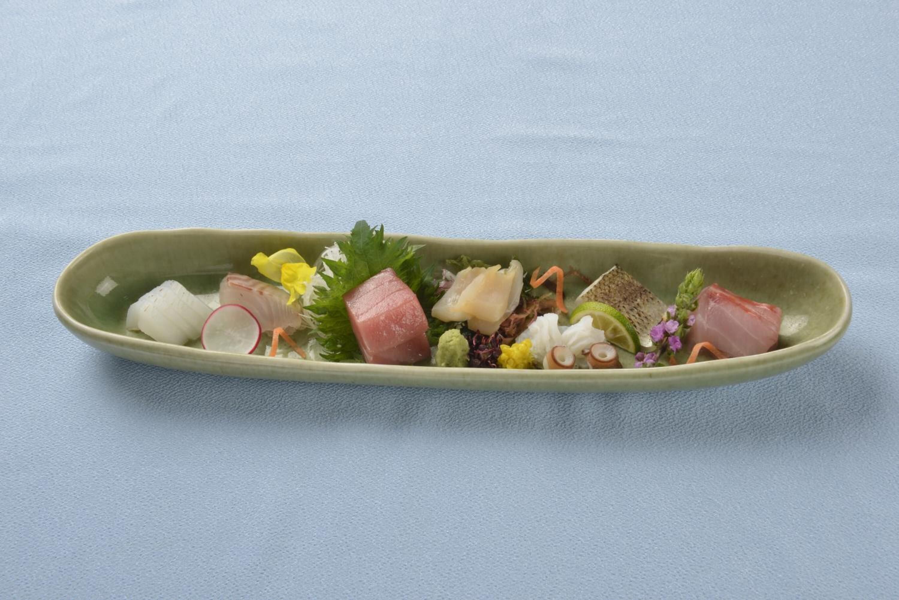 Een rechthoekig keramieken bord met diverse ingrediënten, waaronder vis, groenten en kruiden, op een lichtblauwe achtergrond.