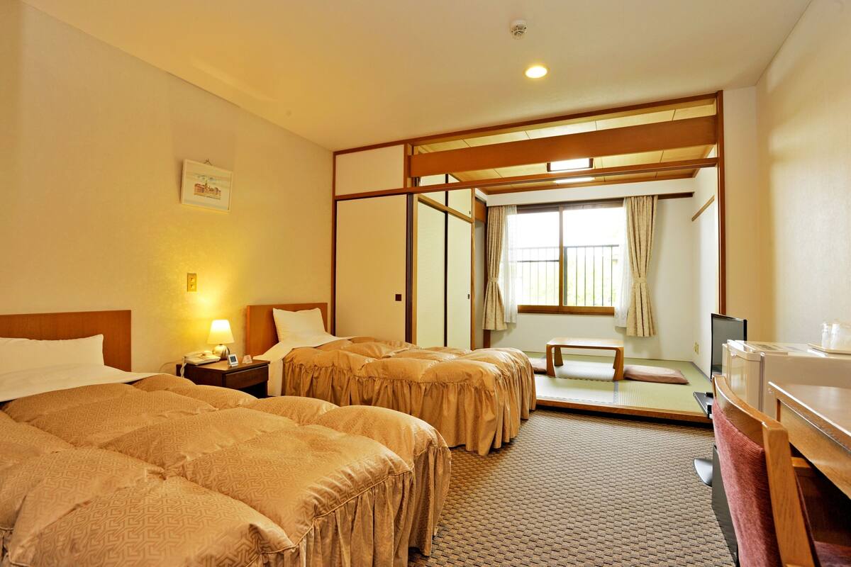 A hotel room with two beds, a desk, a chair, a TV, and a sliding door leading to a balcony.