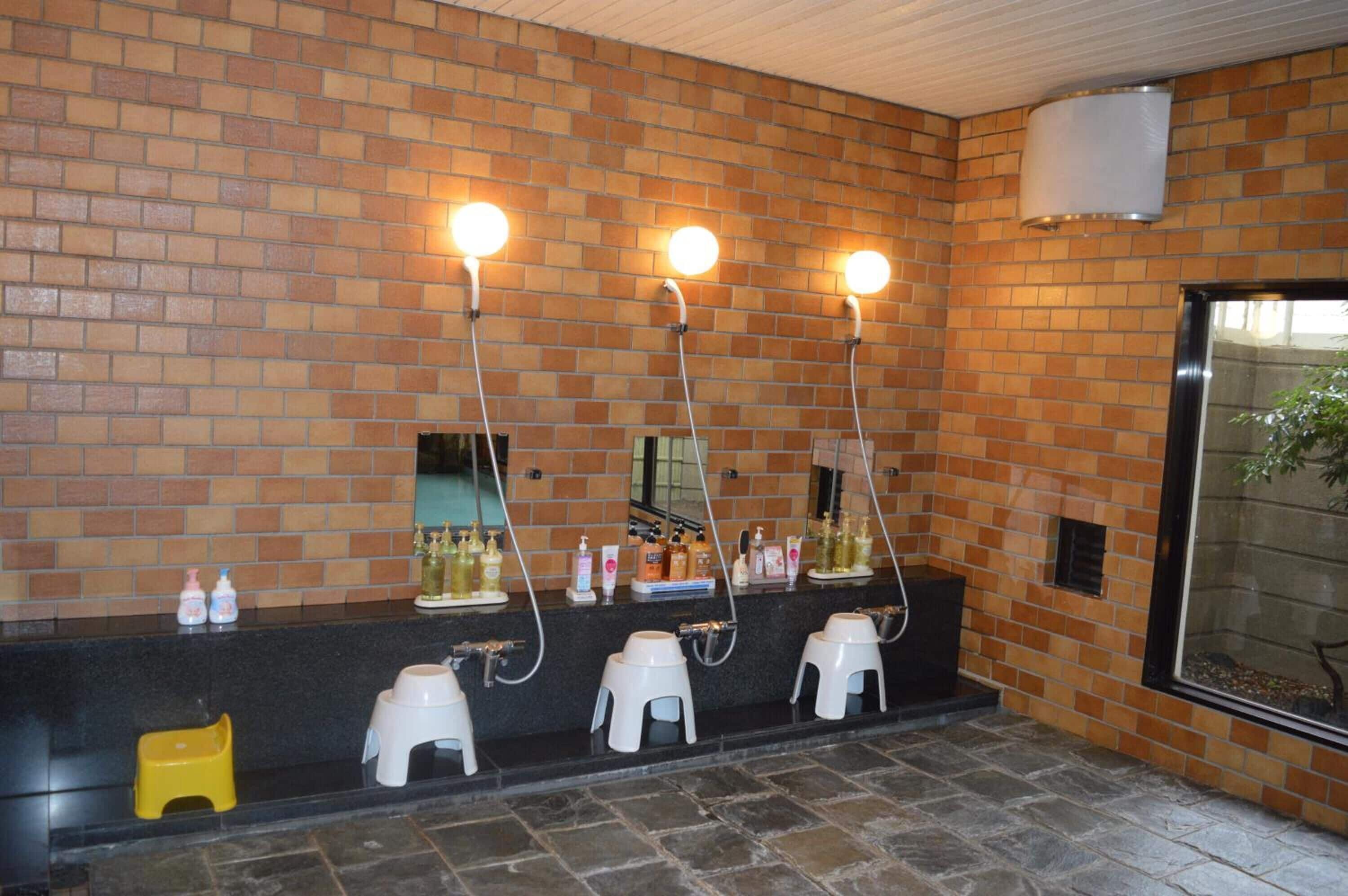 A bathroom with three sinks, each with a mirror and overhead lighting. There are toiletries on the counter, and a window with a view of the outside.