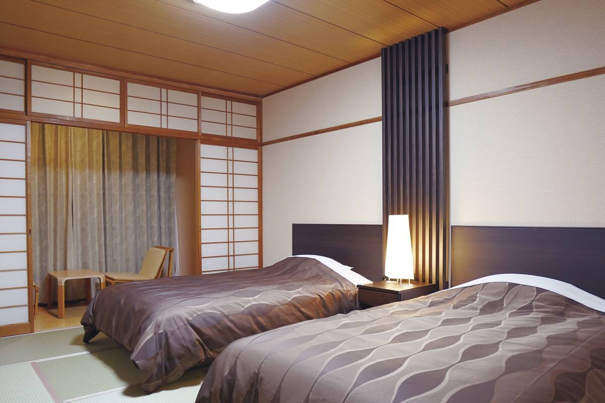 A traditional Japanese room with tatami flooring, two beds with matching bedding, a desk, and a chair.