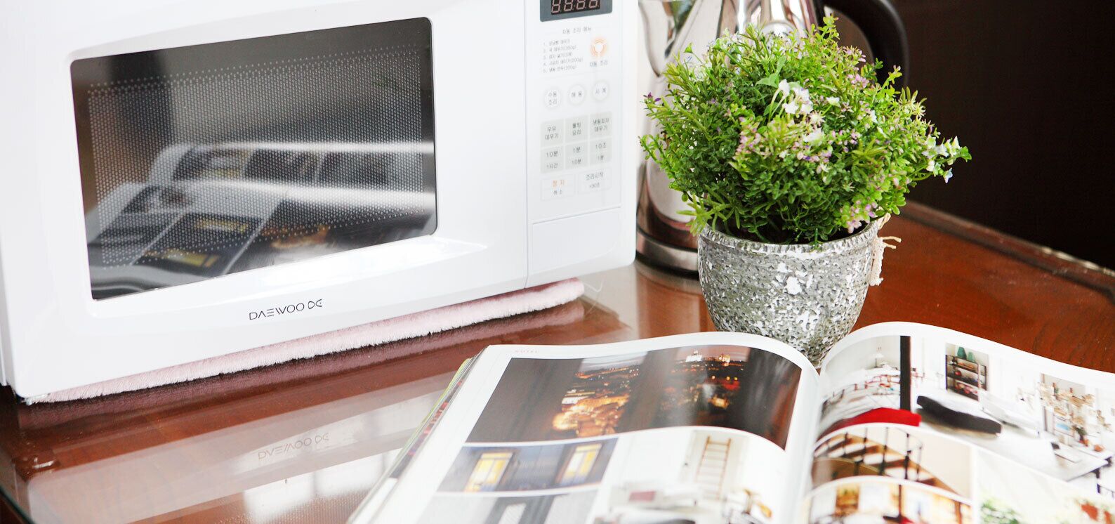 Un microondas blanco con pantalla digital, una pequeña planta en maceta y una revista abierta sobre una mesa de madera.