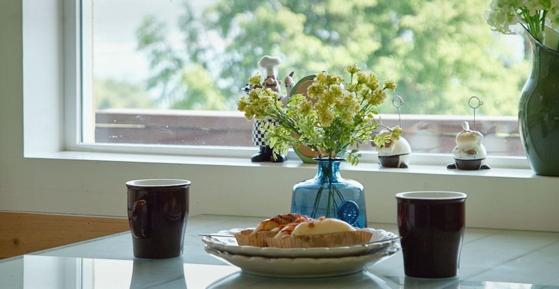 A table with a plate of food, two cups, a vase with flowers, and kitchen items on a windowsill.