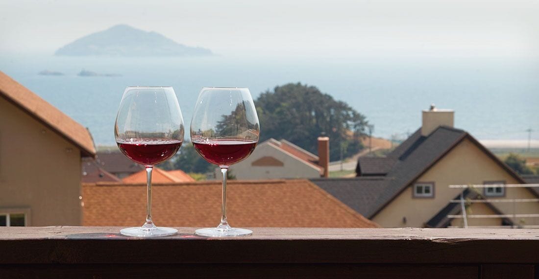 Two wine glasses on a balcony with a view of houses and a distant island.