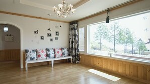 Una habitación con pisos de madera, un banco blanco con cojines florales, una araña y un ventanal con persianas.