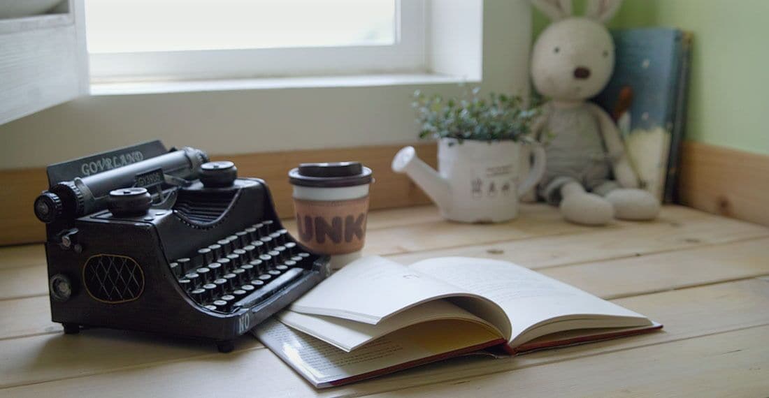 Una máquina de escribir antigua, un libro abierto, una taza con la etiqueta “UNK”, un peluche y una planta en maceta sobre una mesa de madera junto a una ventana.