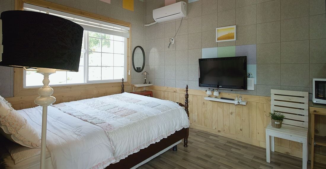 Un dormitorio con cama, televisor, mesita de noche, lámpara y ventana.
