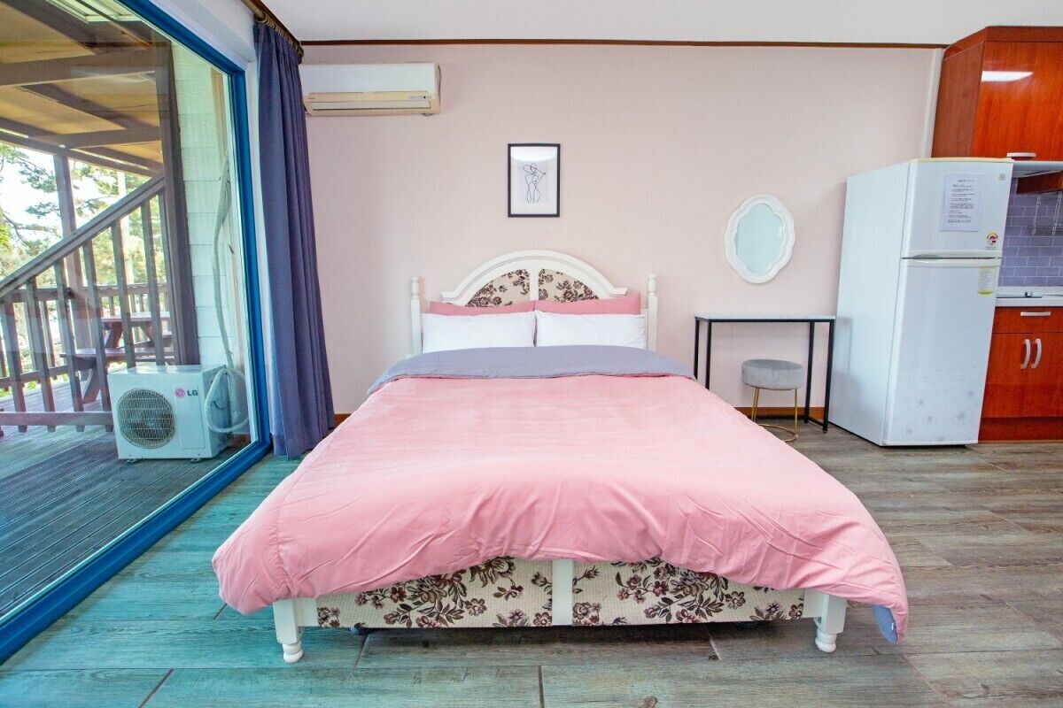 A bedroom with a bed, a small table, a refrigerator, and a window with curtains.
