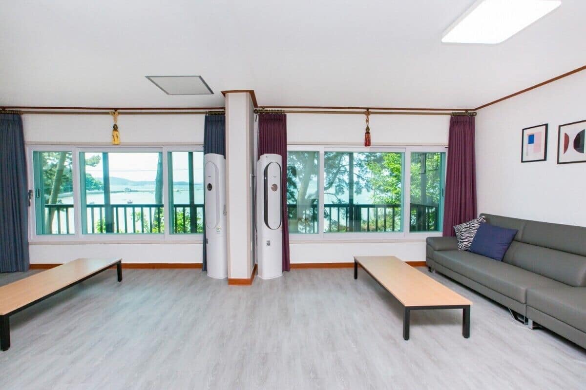 A room with two large windows, a grey sofa, a wooden bench, and two air conditioning units.