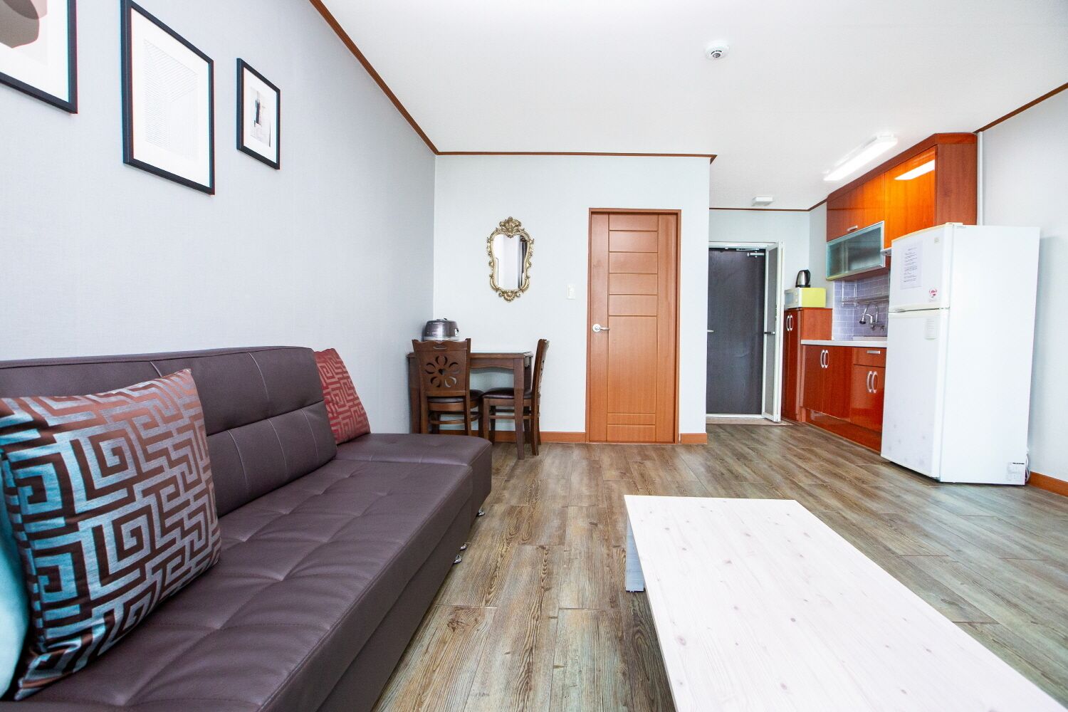 A living room with a sofa, a coffee table, and a dining table with chairs. There is a kitchen area with cabinets and a refrigerator.