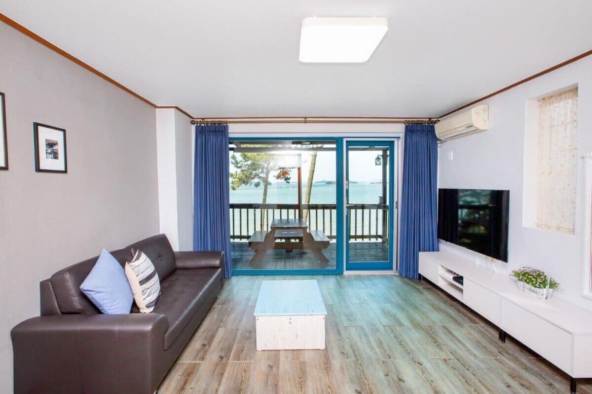 A living room with a sofa, a coffee table, a TV, and a sliding glass door leading to an outdoor area with a view of the sea.