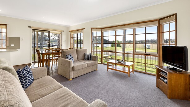 A spacious living room with a large window, a sofa set, a television, and a dining area. - Riviera Beach Resort (Lakes Entrance)