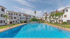 Una piscina rodeada de edificios blancos con balcones, una palmera y un cielo despejado.