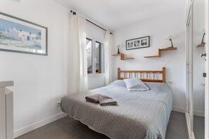 Uma cama bem arrumada com colcha estampada e duas toalhas dobradas, cabeceira de madeira, um quadro emoldurado na parede e uma janela com cortinas.