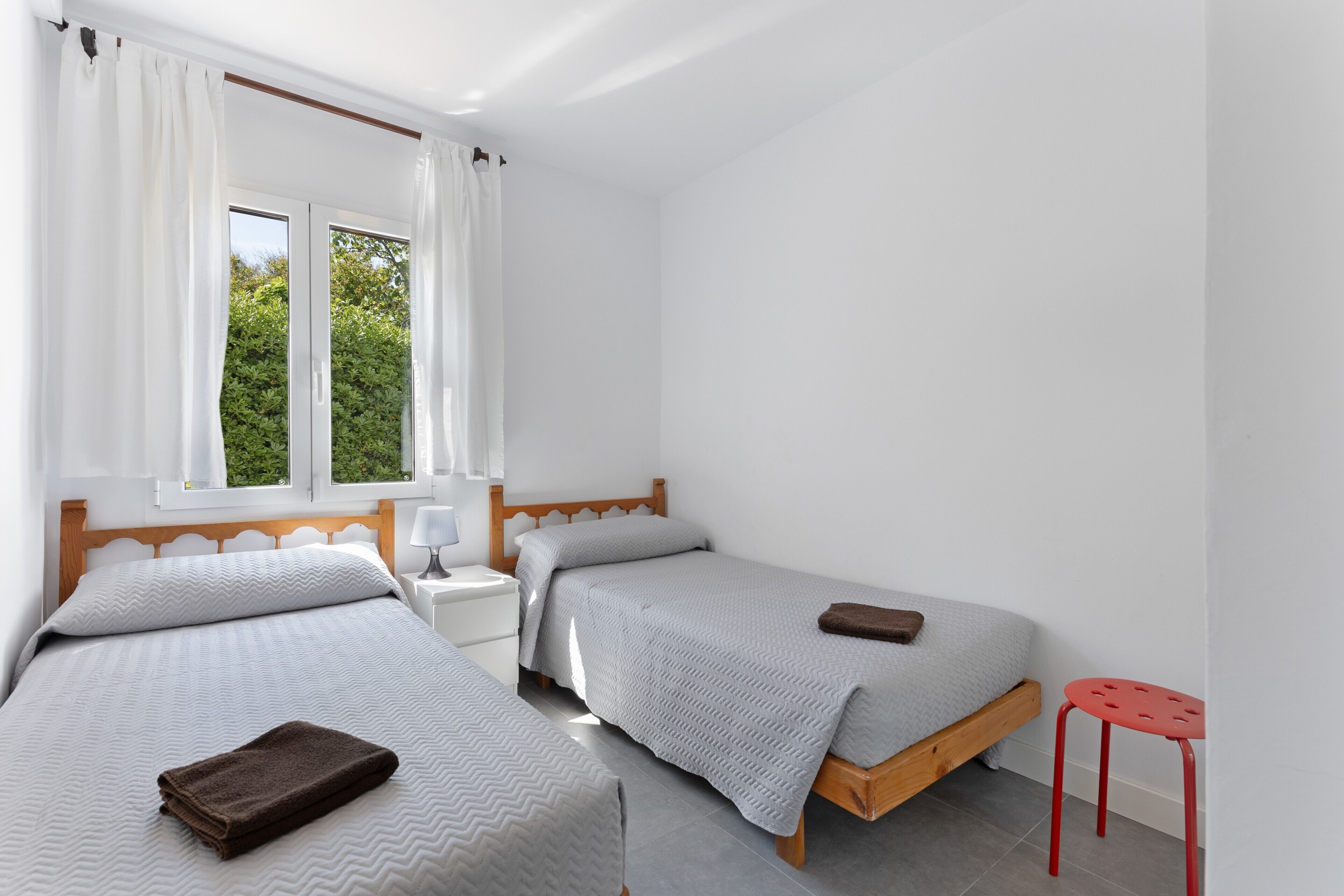 A bedroom with two single beds, a nightstand, a red stool, and a window with curtains.