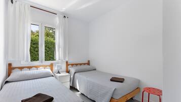 A bedroom with two single beds, a nightstand, a red stool, and a window with curtains.