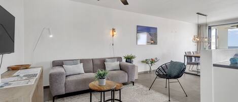 A modern living room with a grey sofa, a wooden coffee table, a black chair, a ceiling fan, and a large window.