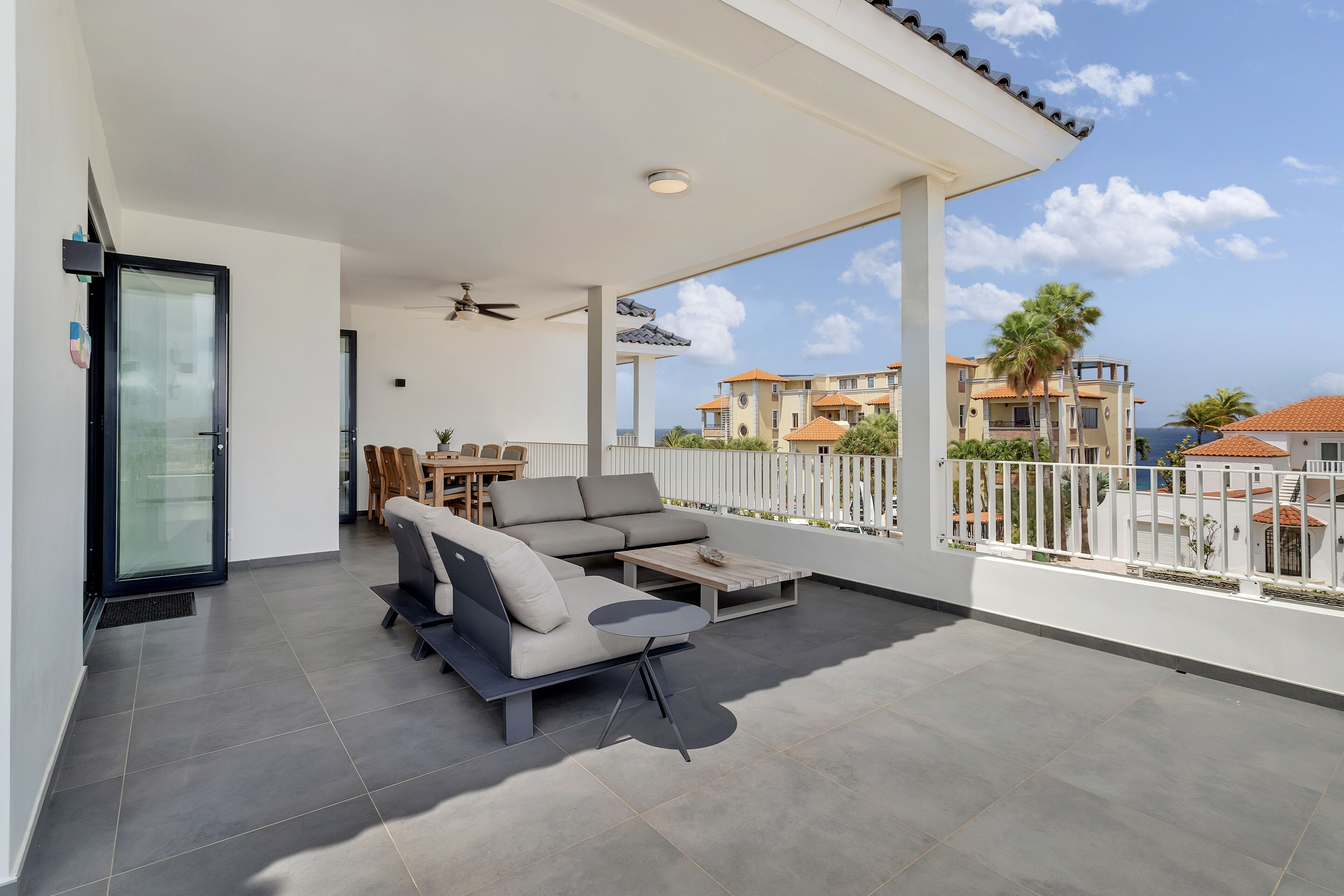 A covered outdoor area with a sofa and table, a glass door, and a view of buildings and palm trees.