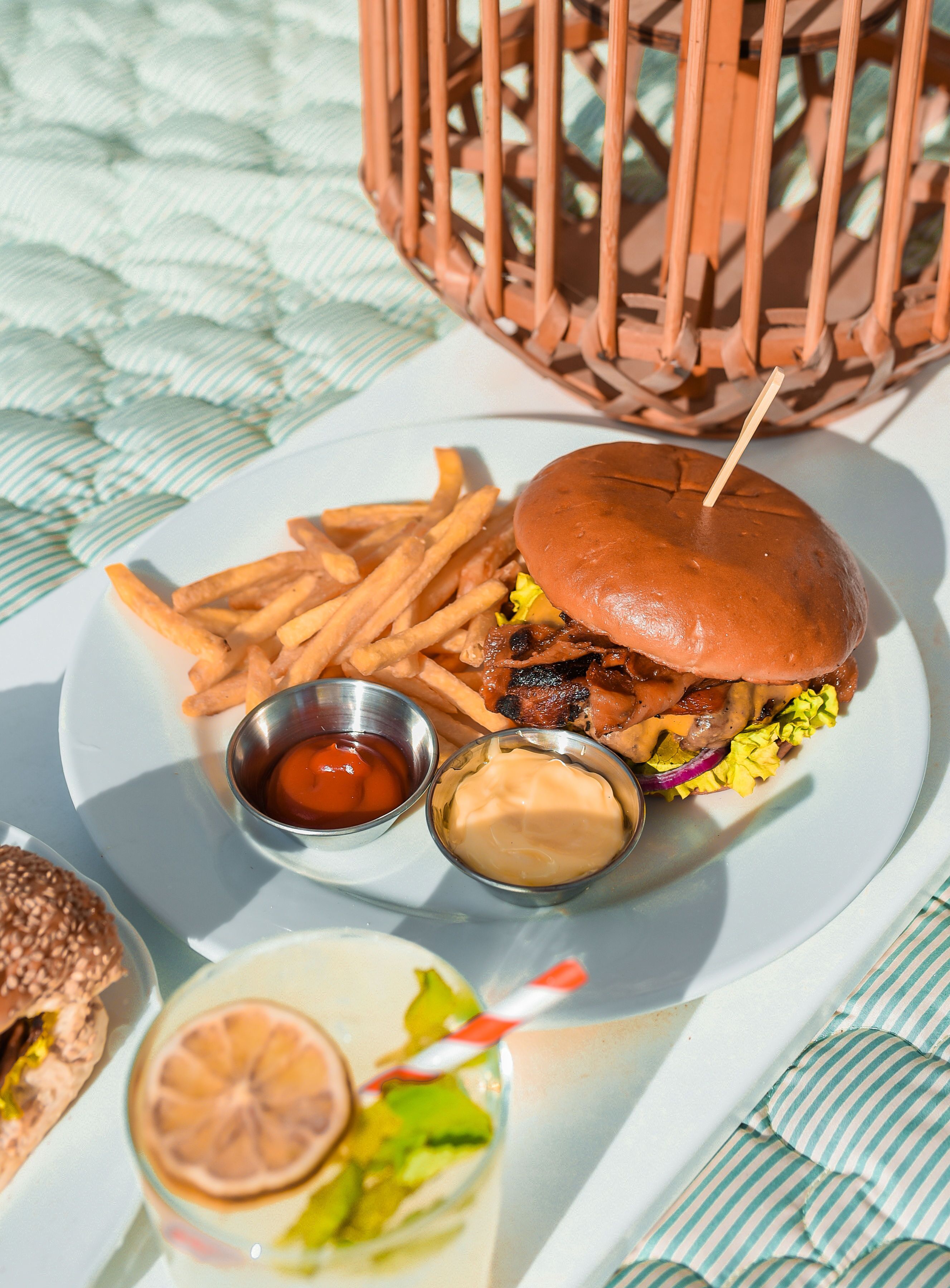 A meal consisting of a burger, fries, and two dipping sauces on a white plate, with a wicker basket and a striped cloth in the background.