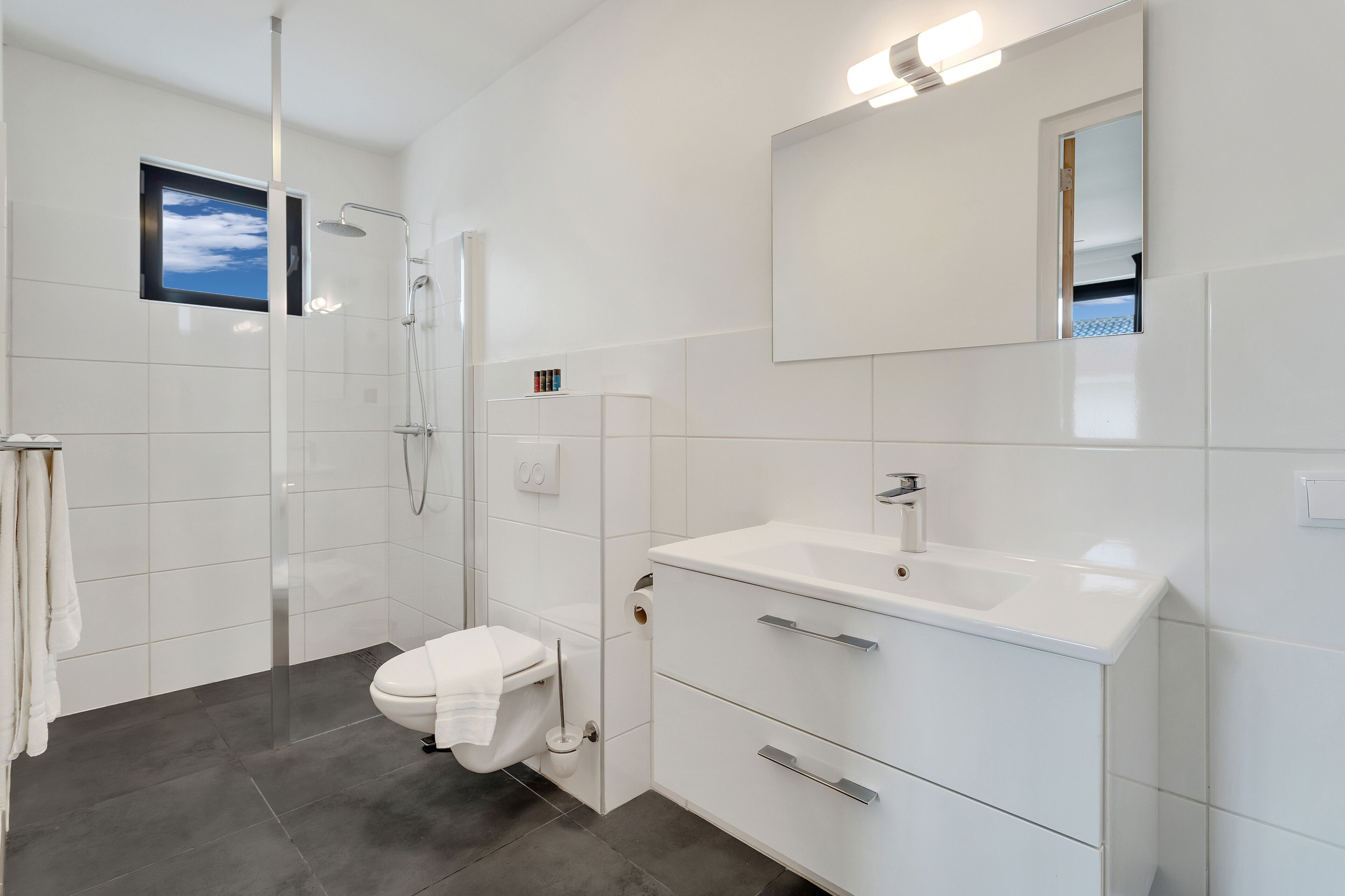 A modern bathroom with a glass shower enclosure, a toilet, a sink with a mirror, and a small window displaying a blue sky.