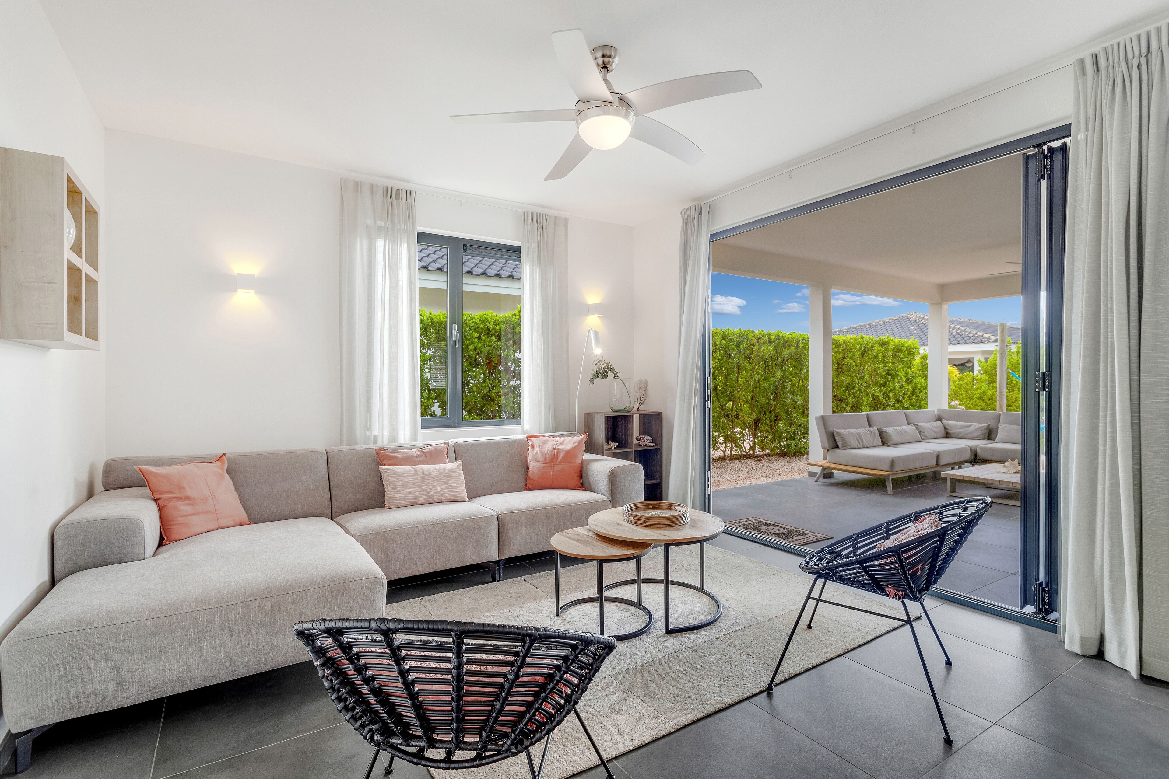Een moderne woonkamer met een bank, twee fauteuils, een plafondventilator en uitzicht door grote schuifdeuren op een terras.