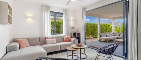 Una sala de estar moderna con un sofá, dos sillones, un ventilador de techo y vistas a un patio exterior a través de amplios puertas correderas.