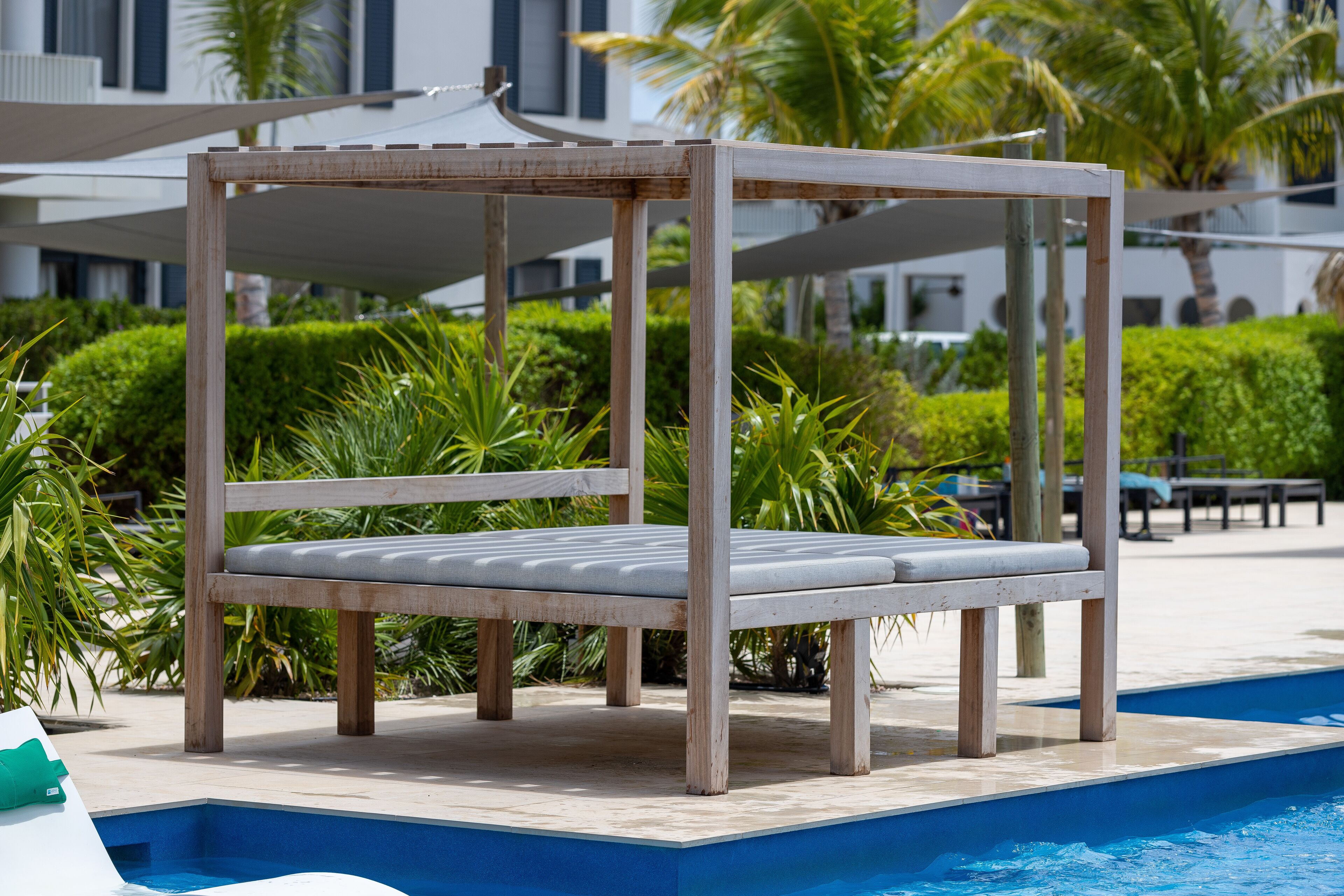 A poolside chair with a wooden frame and grey cushions, positioned near a swimming pool with a clear blue edge.